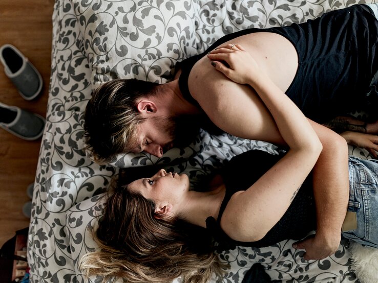 Zwei Personen liegen eng umschlungen auf einem Bett mit floralem Muster, Holzfußboden und grauen Hausschuhen im Hintergrund.