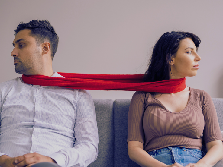Eine Frau in braunem Shirt und ein Mann in weißem Hemd sitzen auf einem grauen Sofa, verbunden durch einen roten Schal.