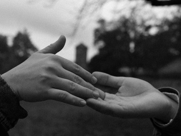 Zwei Hände in einem zarten Berührungsgestus, eine trägt eine Jeansjacke, vor einer parkähnlichen Kulisse in Schwarz-Weiß.