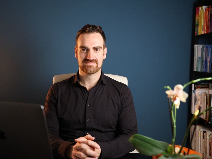Ein Mann in schwarzem Hemd sitzt an einem Schreibtisch, umgeben von einem Laptop, einer weißen Orchidee und Bücherregalen vor einer dunkelblauen Wand. | © Ramón Schlemmbach 