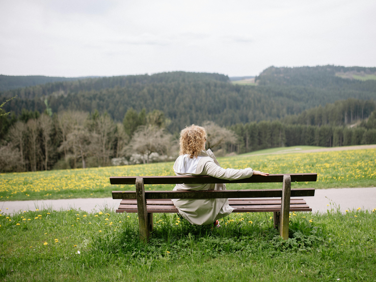 Eine Frau mit lockigem Haar und weißem Mantel sitzt entspannt auf einer Holzbänken, blickt auf eine blühende Frühlingswiese und Waldlandschaft.