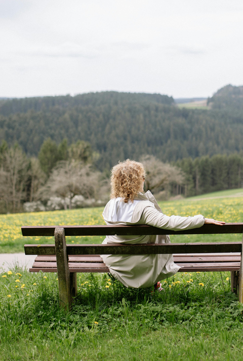 Eine Frau mit lockigem Haar und weißem Mantel sitzt entspannt auf einer Holzbänken, blickt auf eine blühende Frühlingswiese und Waldlandschaft.
