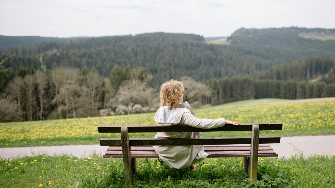 Eine Frau mit lockigem Haar und weißem Mantel sitzt entspannt auf einer Holzbänken, blickt auf eine blühende Frühlingswiese und Waldlandschaft.