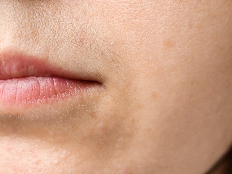 Nahaufnahme des unteren Gesichtsbereichs einer Frau, fokussiert auf die Mundpartie und die Haut darum herum. Die Lippen sind geschlossen und natürlich rosa, und die Haut zeigt Poren und ein paar feine Härchen, wie einen leichten Damenbart, auf der Oberlippe. Es sind auch einige dezente, dunklere Flecken auf der Haut sichtbar, die auf Pigmentierung hinweisen könnten