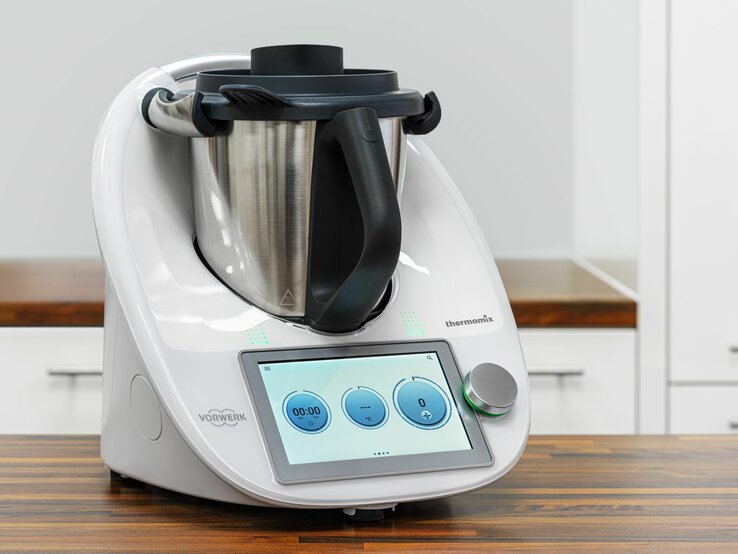 Das Bild zeigt einen modernen Thermomix Küchenhelfer von Vorwerk auf einer Küchenarbeitsplatte. Das Gerät verfügt über ein integriertes digitales Bedienfeld mit einem Touchscreen, auf dem eine Uhr und Bedienelemente zu sehen sind. Der Thermomix besteht aus einer Basisstation mit einem Display und Bedienelementen sowie einem großen Mixtopf aus Edelstahl mit einem schwarzen Griff und einem Messer im Inneren. Auf der rechten Seite des Geräts ist eine Öffnung für den Varoma-Aufsatz oder andere Zubehörteile zu erkennen. Das Design ist schlicht und in einem sauberen Weiß gehalten, mit dem Vorwerk-Logo deutlich sichtbar an der Vorderseite. Der Hintergrund ist schlicht und lenkt nicht von dem Küchengerät ab, was den Fokus auf den Thermomix richtet.