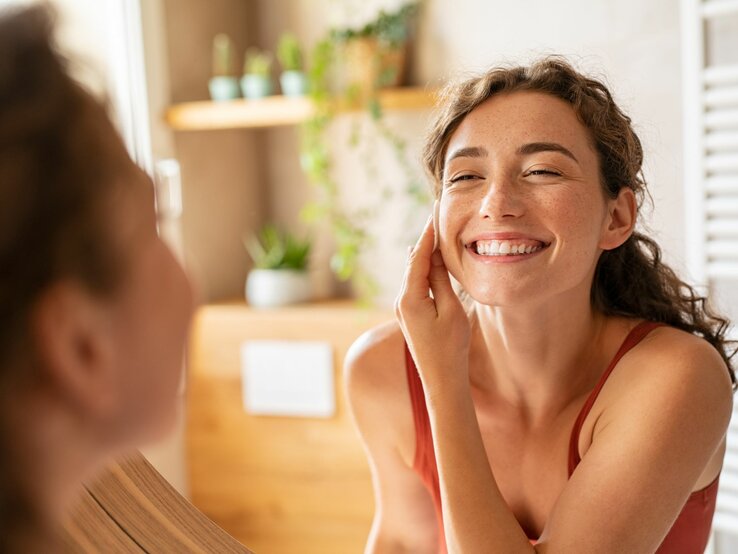 Das Bild zeigt eine junge Frau mit einem herzlichen und offenen Lächeln, die einem anderen Menschen gegenüber sitzt. Die Frau trägt scheinbar keine schwere Make-up-Schicht, aber man kann deutliche, natürlich aussehende Sommersprossen auf ihren Wangen und ihrer Nase erkennen. Diese könnten echte Sommersprossen sein oder sorgfältig mit Make-up geschaffen worden sein, um einen spielerischen und sommerlichen Look zu erzeugen, was darauf hindeutet, dass es sich um den Trend des Schminkens von Fake Sommersprossen handeln könnte. Die Frau hat die Haare locker hochgesteckt und trägt ein einfaches, rotes Tanktop, was den Eindruck eines entspannten und gemütlichen Umfelds verstärkt. Das Umfeld zeigt ein warmes, wohnliches Interieur mit unscharfem Hintergrund, der auf eine gut beleuchtete Küche oder ein Wohnzimmer hindeutet.