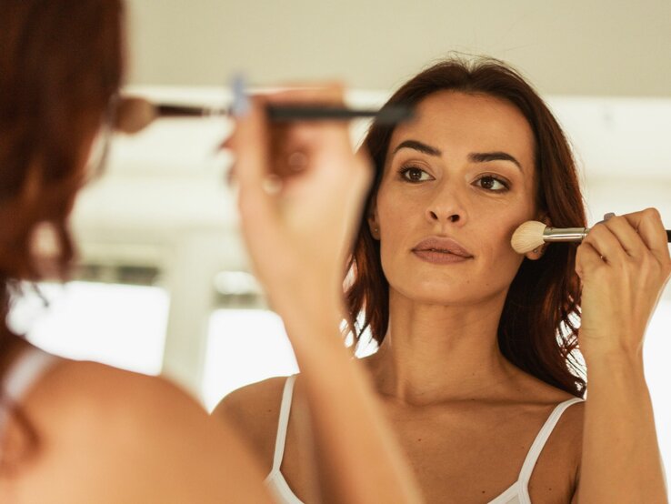 Frau steht vor einem Spiegel und trägt sorgfältig mit einem Kosmetikpinsel Foundation auf ihr Gesicht auf. Sie trägt ein weißes Trägertop.