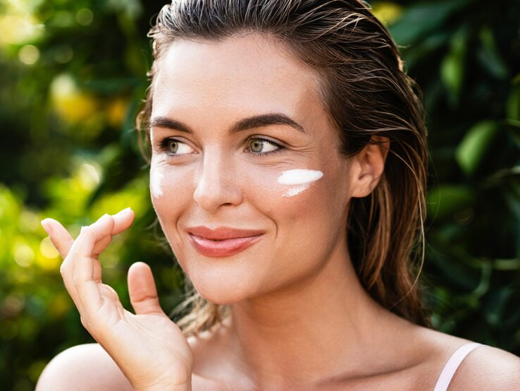 Lächelnde junge Frau mit feuchtem Haar trägt Sonnencreme auf ihre Wange auf, grüne Pflanzen im Hintergrund.