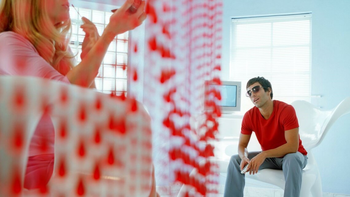 Mann sieht einer Frau beim Schminken zu. Die Frau sitzt links im Bild und hält einen Spiegel, während sie sich schminkt. Der Mann sitzt rechts im Bild auf einem weißen Stuhl, trägt eine Sonnenbrille und ein rotes T-Shirt und schaut die Frau an.