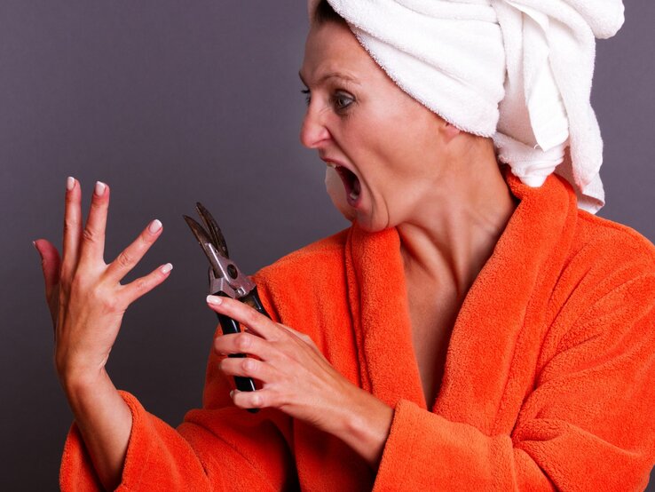 Frau in einem orangefarbenen Bademantel und mit einem weißen Handtuch um den Kopf gewickelt. Sie schaut schockiert auf ihre Hand, während sie eine große Gartenschere hält, als ob sie versucht, ihre Fingernägel damit zu schneiden.