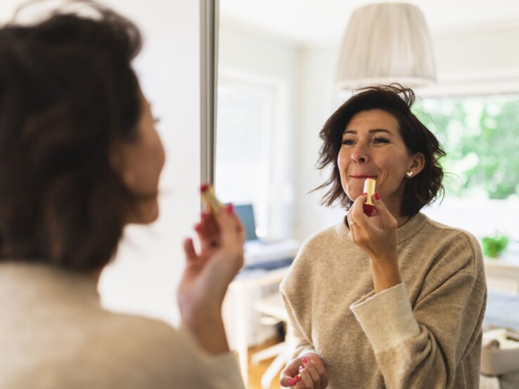 Frau im beigen Pullover trägt Lippenstift vor einem Spiegel auf, im Hintergrund ein gemütliches, gut beleuchtetes Wohnzimmer.