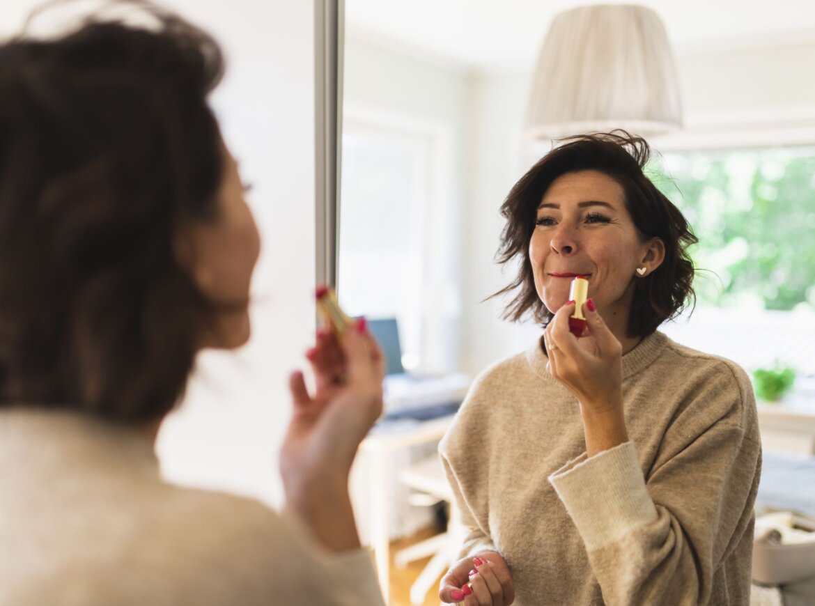 Frau im beigen Pullover trägt Lippenstift vor einem Spiegel auf, im Hintergrund ein gemütliches, gut beleuchtetes Wohnzimmer.