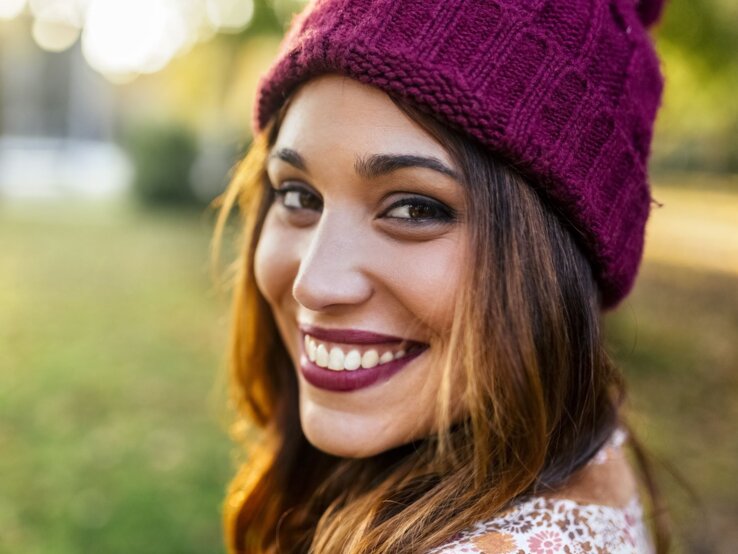 Junge Frau mit lebendigem Lächeln und bordeauxfarbener Strickmütze in herbstlicher Parklandschaft im goldenen Licht.