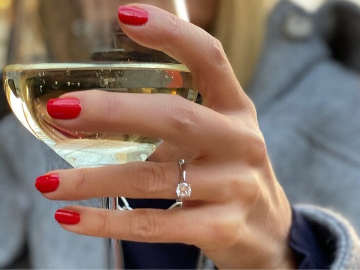 Nahaufnahme einer Hand mit rotem Nagellack, die ein Glas Weißwein hält, Diamantring und grauer Stoff im Hintergrund.