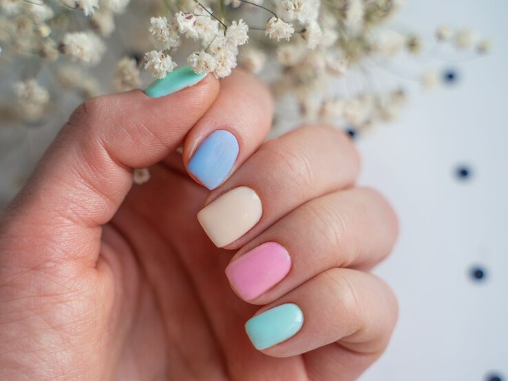 Nahaufnahme einer Hand mit kurzen Nägeln in zartem Pastell-Look, dahinter verschwommene weiße Trockenblumen.