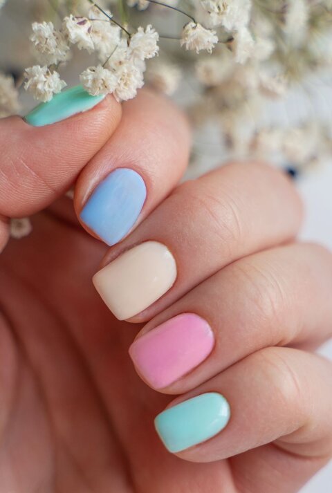 Nahaufnahme einer Hand mit kurzen Nägeln in zartem Pastell-Look, dahinter verschwommene weiße Trockenblumen.