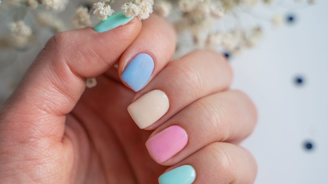 Nahaufnahme einer Hand mit kurzen Nägeln in zartem Pastell-Look, dahinter verschwommene weiße Trockenblumen.