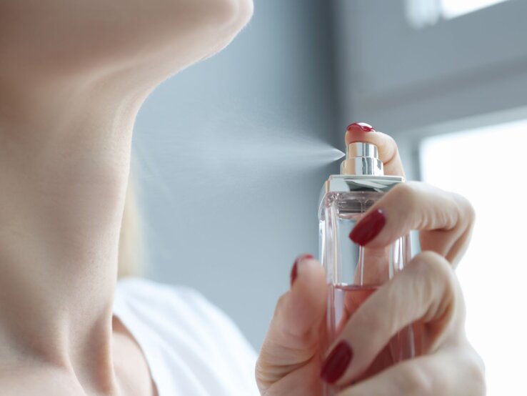 Eine Frau mit roten Nägeln sprüht zarten Parfümnebel auf ihren Hals vor einem hellen Fenster im Morgenlicht.