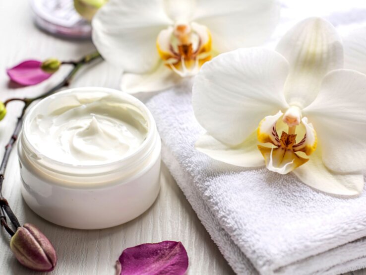 Eine luxuriöse Anti-Aging-Creme in Glasdose liegt auf einem weißen Handtuch, umgeben von Orchideen und Blütenblättern.