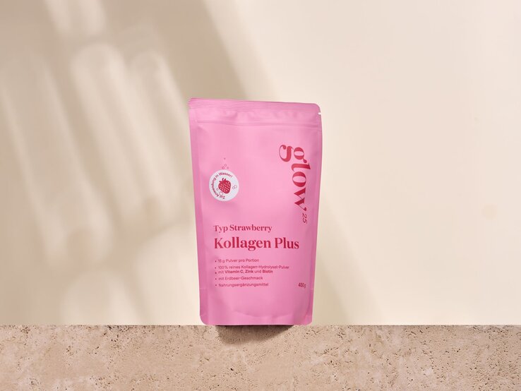 Kollagen Pulver Produktbild | © Glow25 Pinke Verpackung „Kollagen Plus Typ Strawberry“ steht auf beigem Stein vor hellcremer Wand mit weichen Schatten. | © Glow25