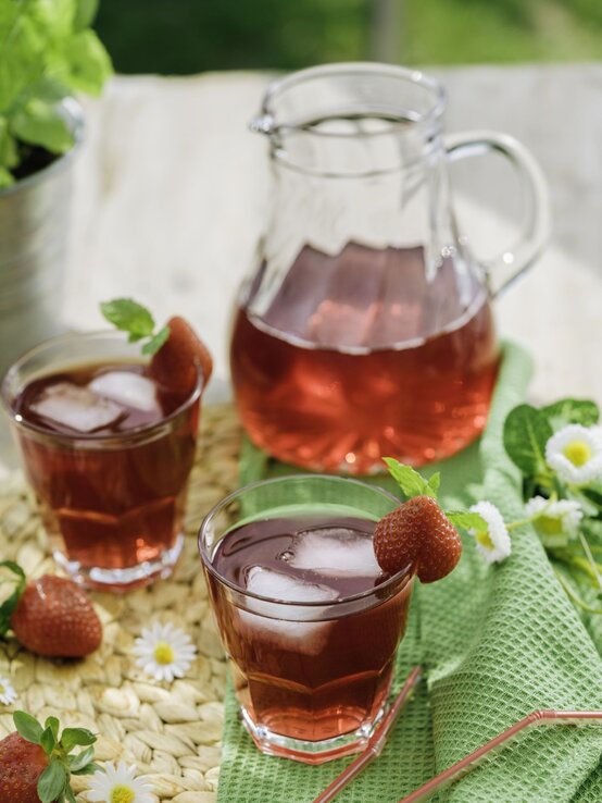 Kollagen Eistee Erdbeere | © Getty Images/ GMVozd Erdbeer-Eistee mit Eis und frischer Deko in Gläsern auf Bastmatte, daneben Gänseblümchen, Minze und eine Karaffe. | © Getty Images/ GMVozd