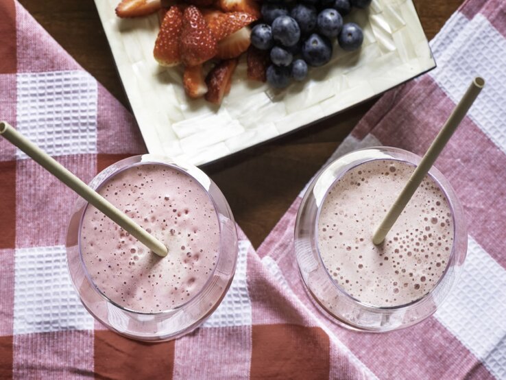 Erdbeer Shake Kollagenpulver | © Getty Images/ Monica Bertolazzi Zwei rosafarbene Smoothies mit Trinkhalm stehen auf rot-weißem Küchentuch, daneben ein Obstteller mit Beerenmix. | © Getty Images/ Monica Bertolazzi