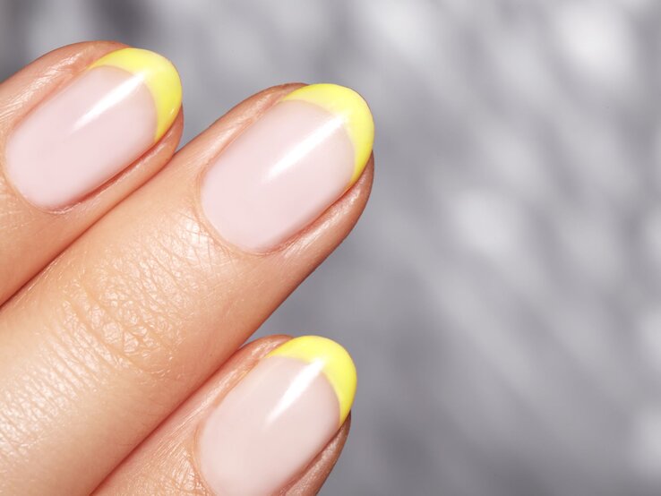 Nahaufnahme einer gepflegten Hand mit pastellrosa Nägeln und leuchtend gelben Spitzen vor einem verschwommenen grauen Hintergrund.