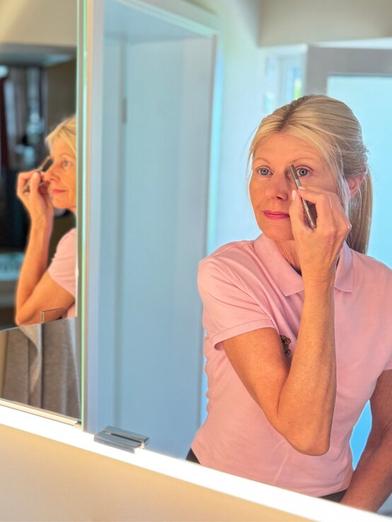 Renate Zott 6 Minuten Make-up Augenbrauen im Spiegel | © Renate Zott Eine schlanke Frau mit blondem Pferdeschwanz trägt ein rosafarbenes Polohemd und schminkt sich vor einem Badezimmer-Spiegel. | © Renate Zott