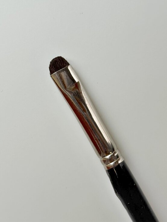 Renate Zott 6 Minuten Make-up Eye Brush | © Renate Zott Ein kleiner, abgerundeter Schminkpinsel mit schwarzem Griff und silberner Metallfassung liegt auf einer weißen Oberfläche. | © Renate Zott