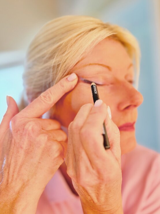 Renate Zott 6 Minuten Make-up Kajal | © Renate Zott Eine Frau mit blondem Haar deutet mit den Fingern auf ihr Auge und trägt ein rosafarbenes Oberteil in hellem Raum. | © Renate Zott