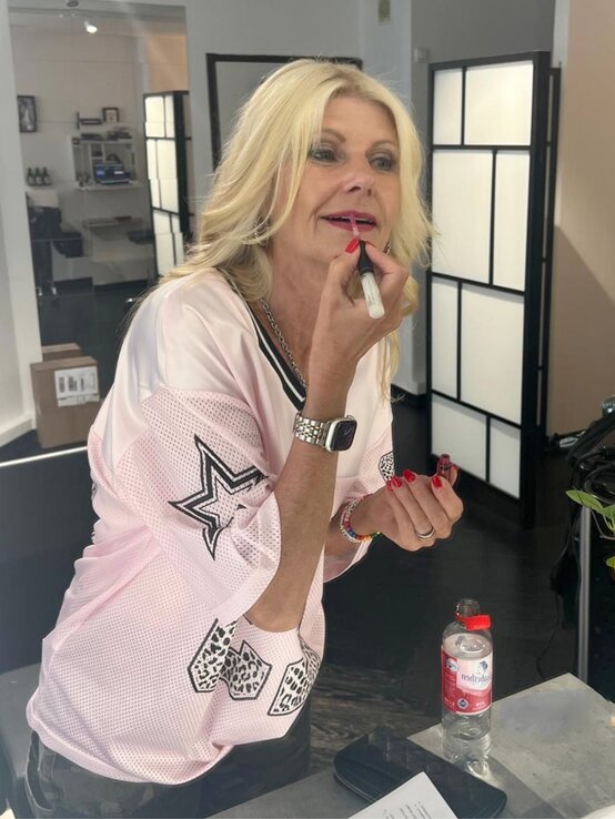 Renate Zott 6 Minuten Make-up Lippenstift | © Renate Zott Eine Frau mit welligem, blondem Haar trägt ein rosafarbenes Oberteil mit schwarzen Sternmotiven und schminkt sich in einem modernen Raum | © Renate Zott