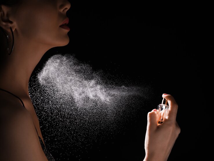 Parfum richtig auftragen Eine weibliche Hand sprüht feinen Parfümnebel aus einem Glasflakon auf einen Hals vor schwarzem Hintergrund.