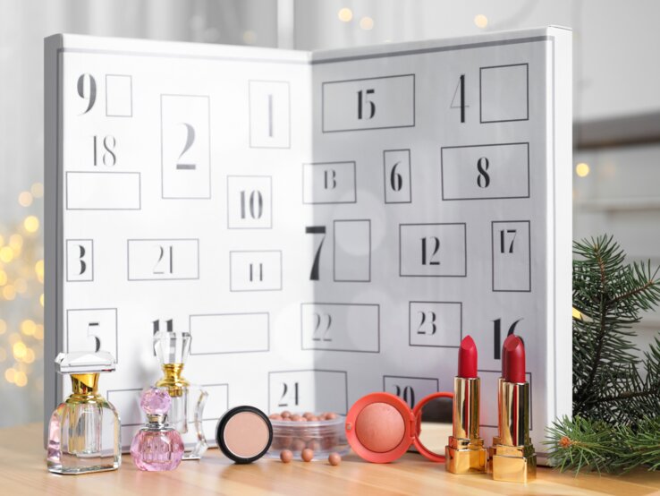 Geöffneter weißer Adventskalender mit nummerierten Türchen, davor stehen Parfümflakons, roter Lippenstift, Rouge und Tannenzweige.