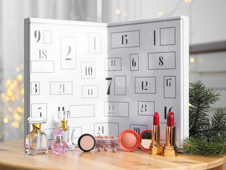 Geöffneter weißer Adventskalender mit nummerierten Türchen, davor stehen Parfümflakons, roter Lippenstift, Rouge und Tannenzweige.