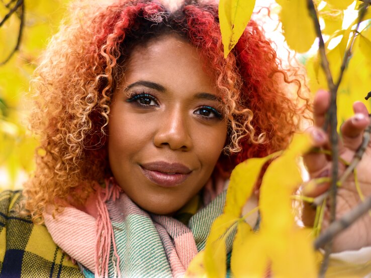 Eine Person mit lockigem, rotem Haar im Fokus, gelbes Herbstlaub im Vordergrund, trägt einen karierten Mantel und pastellfarbenen Schal.