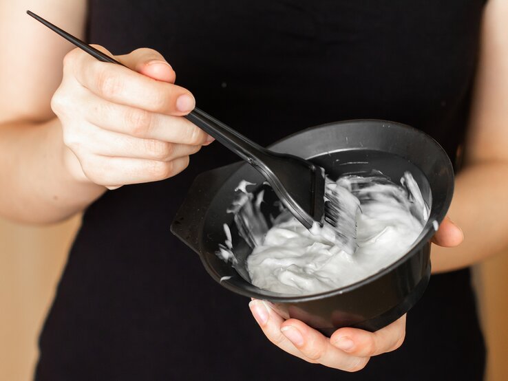 Hände einer Frau halten eine schwarze Schale mit cremiger Haarfarbe, während sie die Farbe mit einem schwarzen Pinsel rührt.