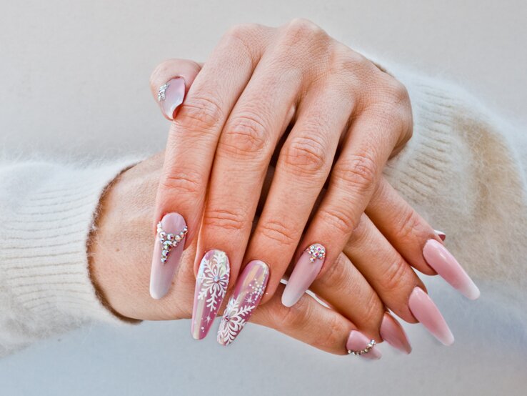 Nahaufnahme von gepflegten Händen mit langen, rosa-weißen Nägeln, verziert mit Glitzersteinen und Schneeflockenmuster vor hellem Hintergrund.