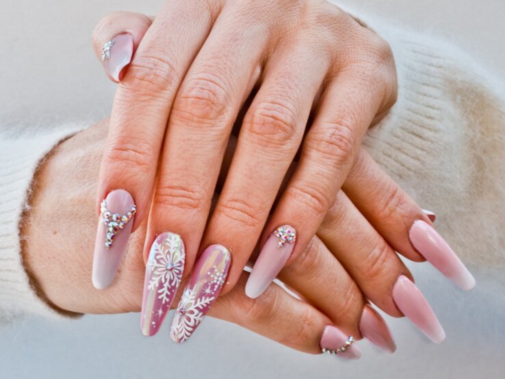 Nahaufnahme von gepflegten Händen mit langen, rosa-weißen Nägeln, verziert mit Glitzersteinen und Schneeflockenmuster vor hellem Hintergrund.
