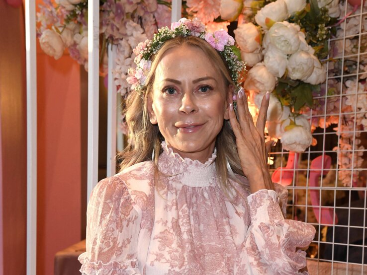 Frau in einem langärmligen, rosa-weiß gemusterten Kleid mit Blumenkranz sitzt vor einer Dekoration mit weißen und rosa Blumen.