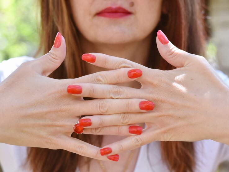 Zwei Hände mit leuchtend roten, lackierten Nägeln und einem roten Ring sind ineinander verschränkt, vor einem unscharfen grünen Hintergrund.