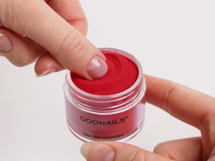 Nahaufnahme eines Fingers bei der Anwendung von rotem Dipping Puder aus einer Glasdose mit minimalistischer Aufschrift. | © Doonails