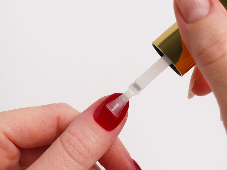 Detailaufnahme des Auftragens eines Klarlacks auf einen tiefrot Dipping Powder Nagel, gehalten von gepflegten Händen. | © Doonails