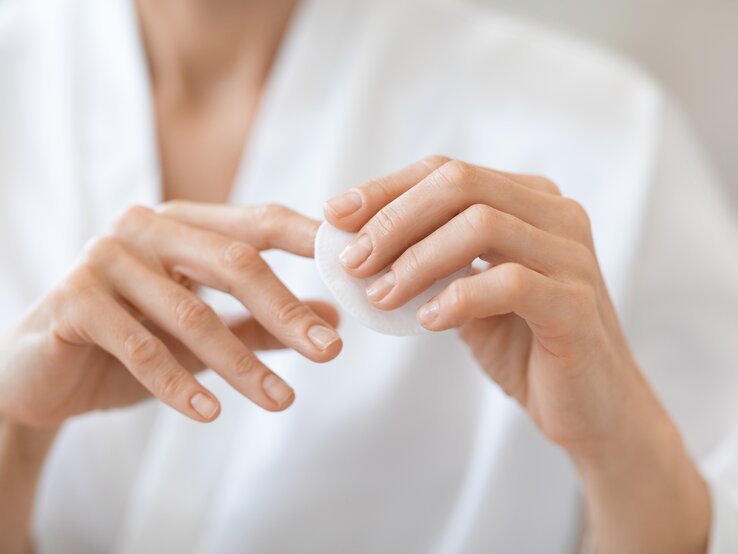 Hände einer Person, die eine weiße Wattepadscheibe zwischen Daumen und Zeigefinger hält, unscharfer Oberkörper in weißem Hemd im Hintergrund.