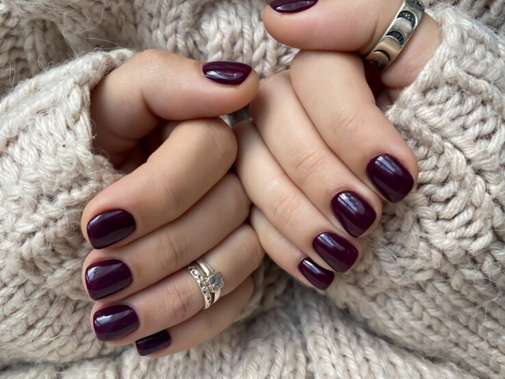 Zwei gepflegte Hände mit dunkelviolett lackierten Nägeln halten sich warm im Ärmel eines beigen Strickpullovers.