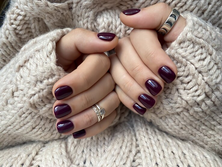 Zwei gepflegte Hände mit dunkelviolett lackierten Nägeln halten sich warm im Ärmel eines beigen Strickpullovers.