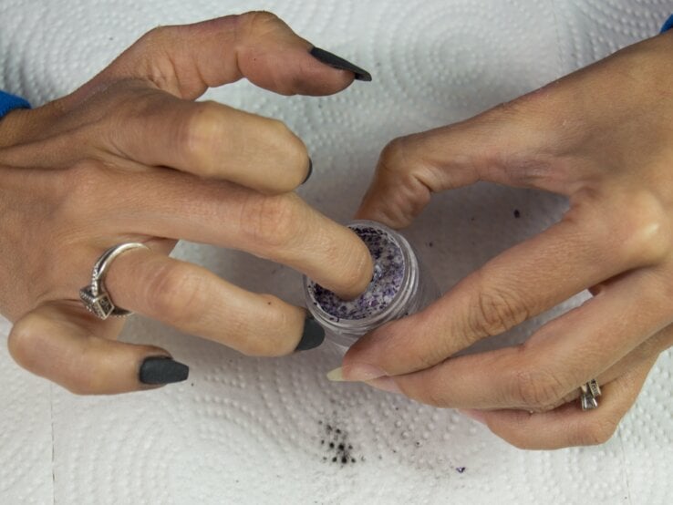 Hände mit dunklem Nagellack, eine Hand taucht Finger in ein kleines Gefäß mit violettem Puder, beide Hände tragen Ringe.