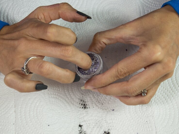 Hände mit dunklem Nagellack, eine Hand taucht Finger in ein kleines Gefäß mit violettem Puder, beide Hände tragen Ringe.
