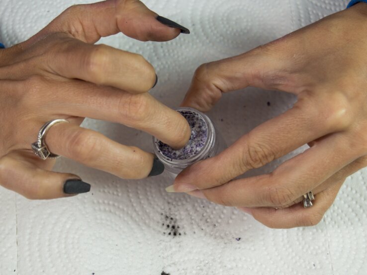 Hände mit dunklem Nagellack, eine Hand taucht Finger in ein kleines Gefäß mit violettem Puder, beide Hände tragen Ringe.