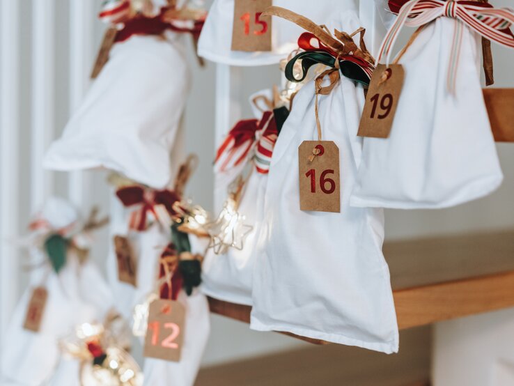 Weiße Stoffbeutel mit roten Zahlen und Schleifen hängen als dekorativer Adventskalender an einer Holztreppe, sanft beleuchtete Sterne.