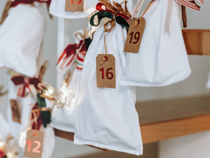Weiße Stoffbeutel mit roten Zahlen und Schleifen hängen als dekorativer Adventskalender an einer Holztreppe, sanft beleuchtete Sterne.